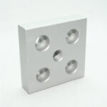 Square aluminum block with four circular holes and a central threaded hole, arranged in a 2x2 plus center pattern.