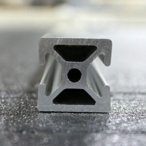 Close-up of a gray aluminum extrusion with an I-beam cross-section resting on a textured workshop floor, showing hollow chambers and a central hole.