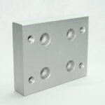 Rectangular silver aluminum mounting plate with six circular holes: four larger through-holes and two smaller threaded holes arranged in a grid, likely used as a fixture base.