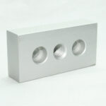 Rectangular gray metal block with three circular holes, the middle hole is threaded.