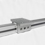 Close-up of a gray industrial linear bearing block mounted on a metal rail with multiple screw holes, set against a light background.