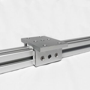 Close-up of a gray industrial linear bearing block mounted on a metal rail with multiple screw holes, set against a light background.